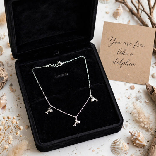 A silver-toned necklace with three small dolphin charms is displayed in an open black velvet jewelry box. The box is styled on a light surface with seashells, dried grass, twigs, and a kraft card that reads, 'You are free like a dolphin.