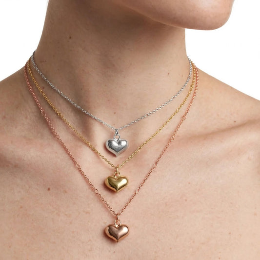 Three delicate heart-shaped pendant necklaces in silver, gold, and rose gold tones layered on a model's neck against a white background.