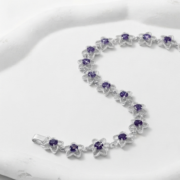 A silver-toned flower-link bracelet with small purple gemstone centers, arranged in an S on a white sculptural tray against a white background.