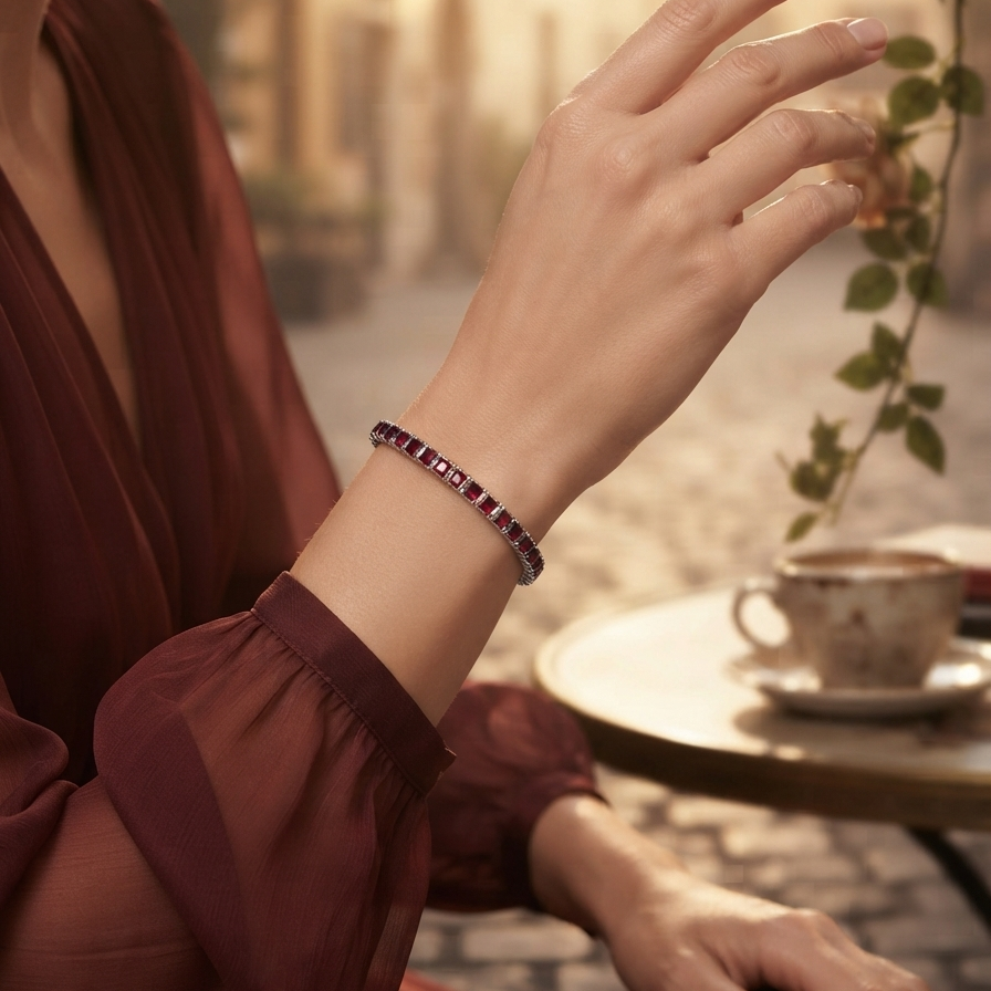 A woman in a burgundy dress wearing a thin bracelet with dark red rectangular stones, posed in a warm, softly lit cafe setting with a cup on a table.