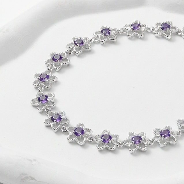 A silver-toned flower-link bracelet with small purple gemstones, displayed on a white sculptural tray against a clean white background.