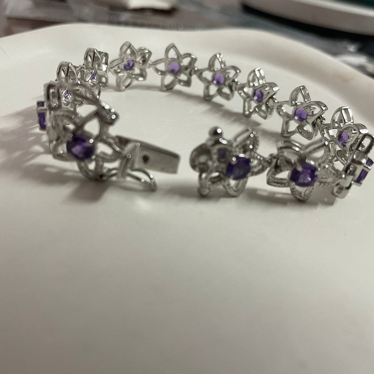 A silver-tone bracelet with repeating flower motifs and small purple gemstones, photographed on a white surface with a blurred indoor background.