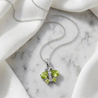 A silver chain necklace with a butterfly pendant featuring two green gemstones and small clear accent stones, styled on draped white fabric over a light marble surface.