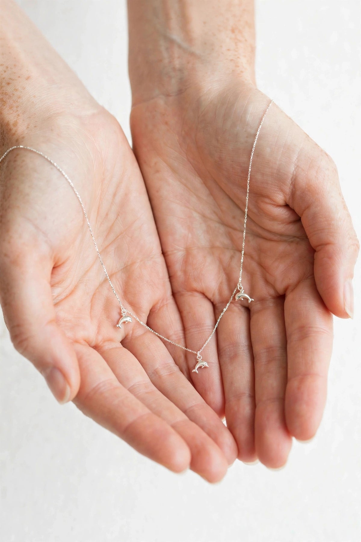 A delicate silver necklace with three small bird charms is draped across two cupped hands against a soft, light off-white background.