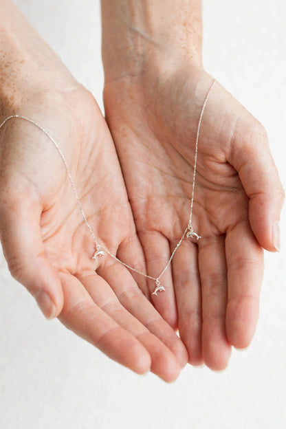 A delicate silver necklace with three small bird charms is draped across two cupped hands against a soft, light off-white background.