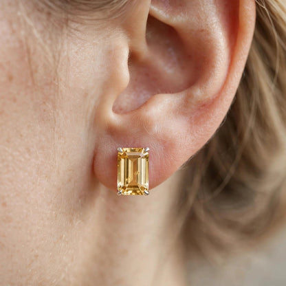 A close-up of a person's ear wearing a small rectangular yellow gemstone stud earring in a gold setting.