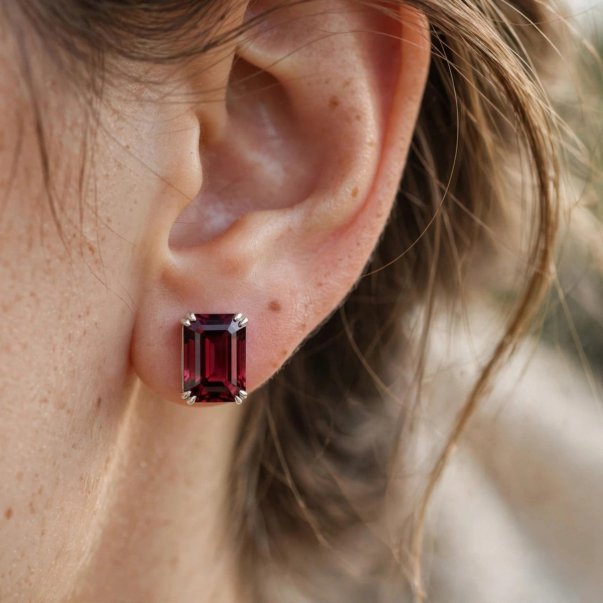 A close-up of a person's ear wearing a rectangular emerald-cut dark red gemstone stud earring in a silver-toned setting.