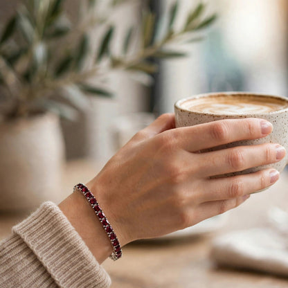 A close-up lifestyle shot of a person's hand holding a ceramic coffee mug with a latte, wearing a dark red beaded bracelet on the wrist. The background is a softly blurred cafe setting with a table, napkin, and potted plant.