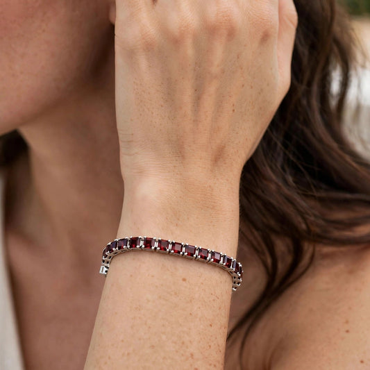 A close-up lifestyle shot of a woman wearing a silver-toned tennis bracelet with small rectangular dark red gemstones, with her hand resting near her face.