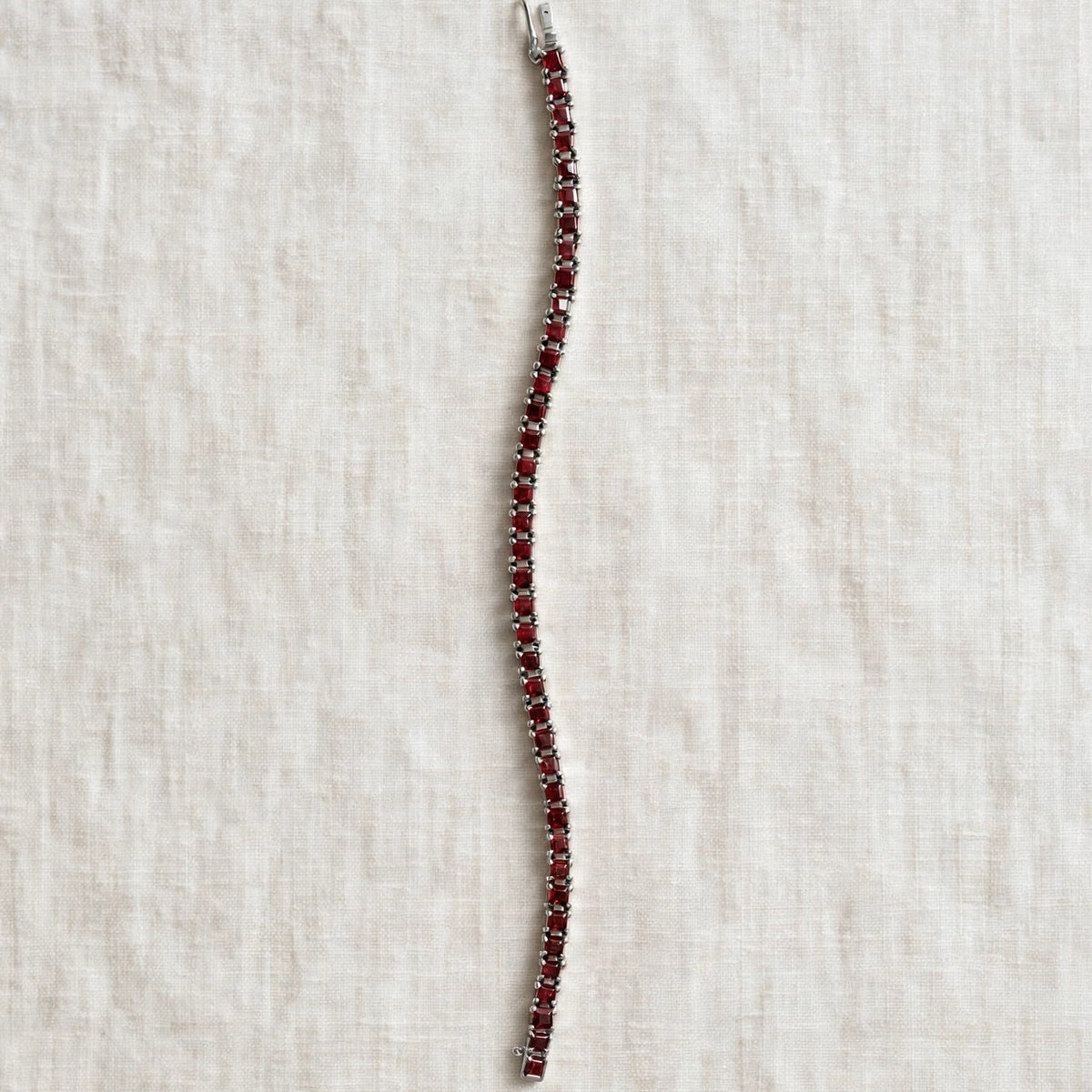 A single-strand tennis bracelet featuring deep red gemstones in a silver-toned setting, laid flat on a textured off-white linen background.