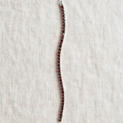 A single-strand tennis bracelet featuring deep red gemstones in a silver-toned setting, laid flat on a textured off-white linen background.