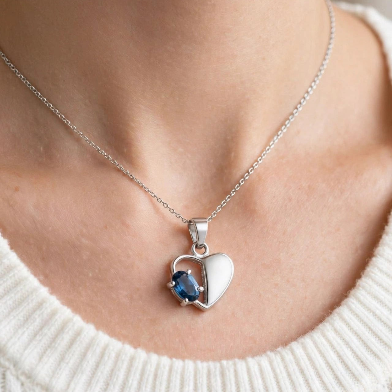 A close-up of a person wearing a silver heart pendant necklace with a split design, one half white and the other half featuring a small blue gemstone, paired with a white ribbed knit sweater.
