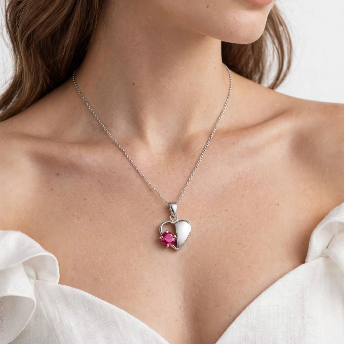 A close-up of a woman wearing a delicate silver heart pendant necklace with a small pink gemstone accent, paired with a white off-the-shoulder blouse against a clean white background.