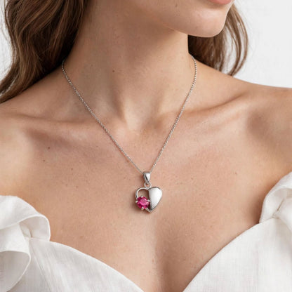 A close-up of a woman wearing a delicate silver heart pendant necklace with a small pink gemstone accent, paired with a white off-the-shoulder blouse against a clean white background.