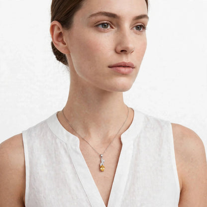 A woman wearing a delicate silver-toned necklace with a pear-cut yellow gemstone pendant, styled with a white linen top against a clean white background.