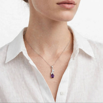 A close-up of a woman wearing a delicate silver-toned necklace with a teardrop pendant featuring a deep purple gemstone and small clear stones, paired with a white collared shirt against a clean white background.