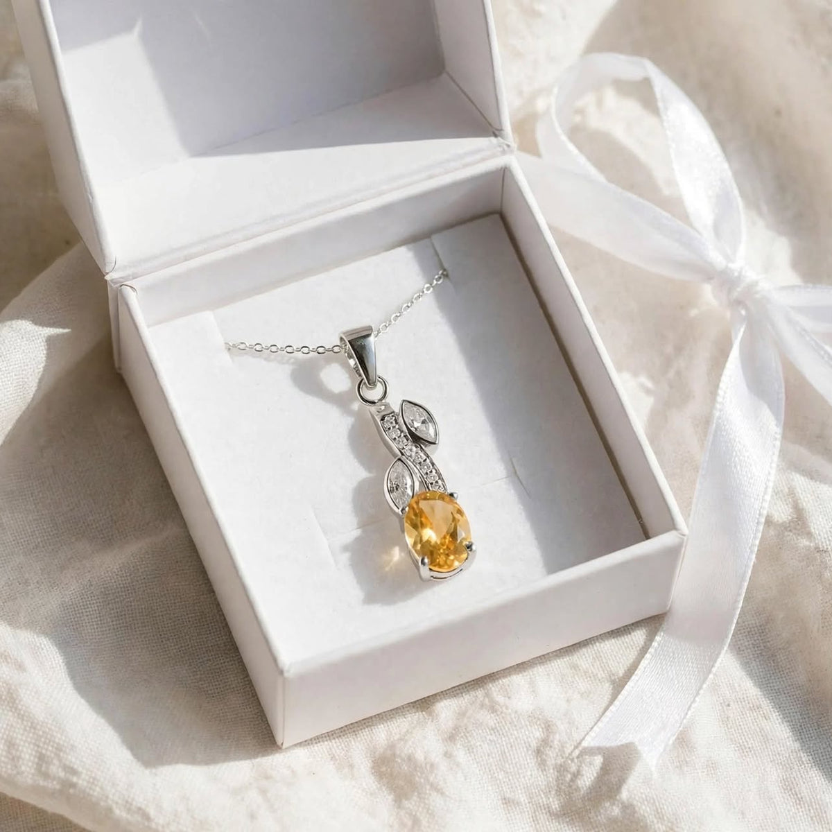 A silver pendant necklace with a yellow gemstone displayed inside an open white jewelry box on a soft beige fabric background with a white ribbon.