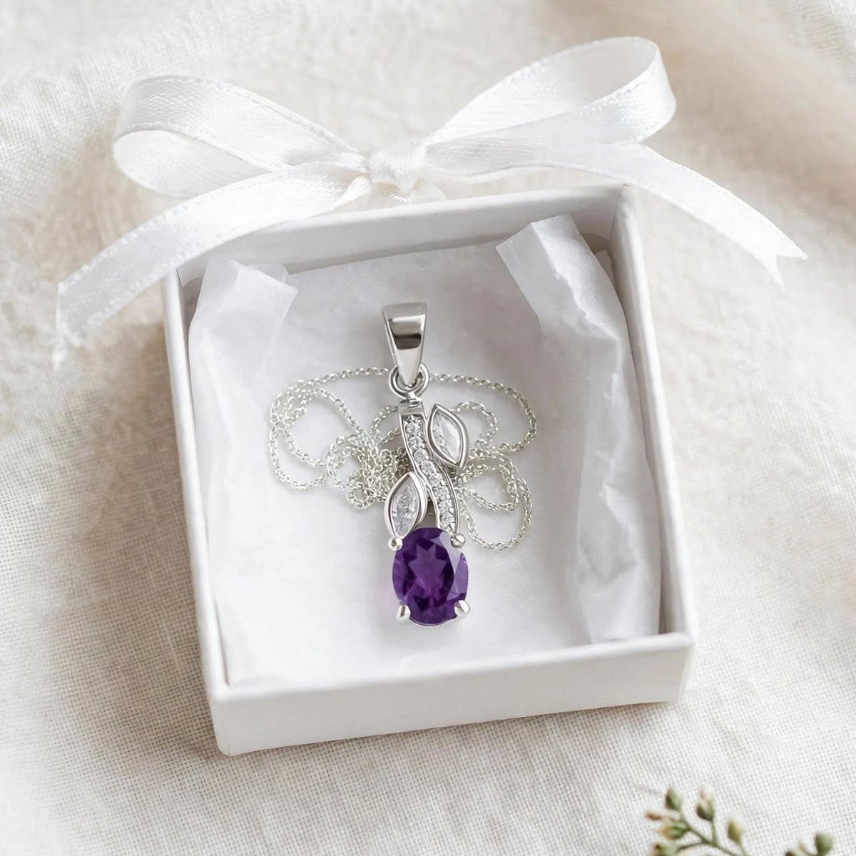 A silver pendant necklace with a teardrop-shaped purple gemstone, presented in a white gift box with a white satin ribbon on a soft white fabric background.