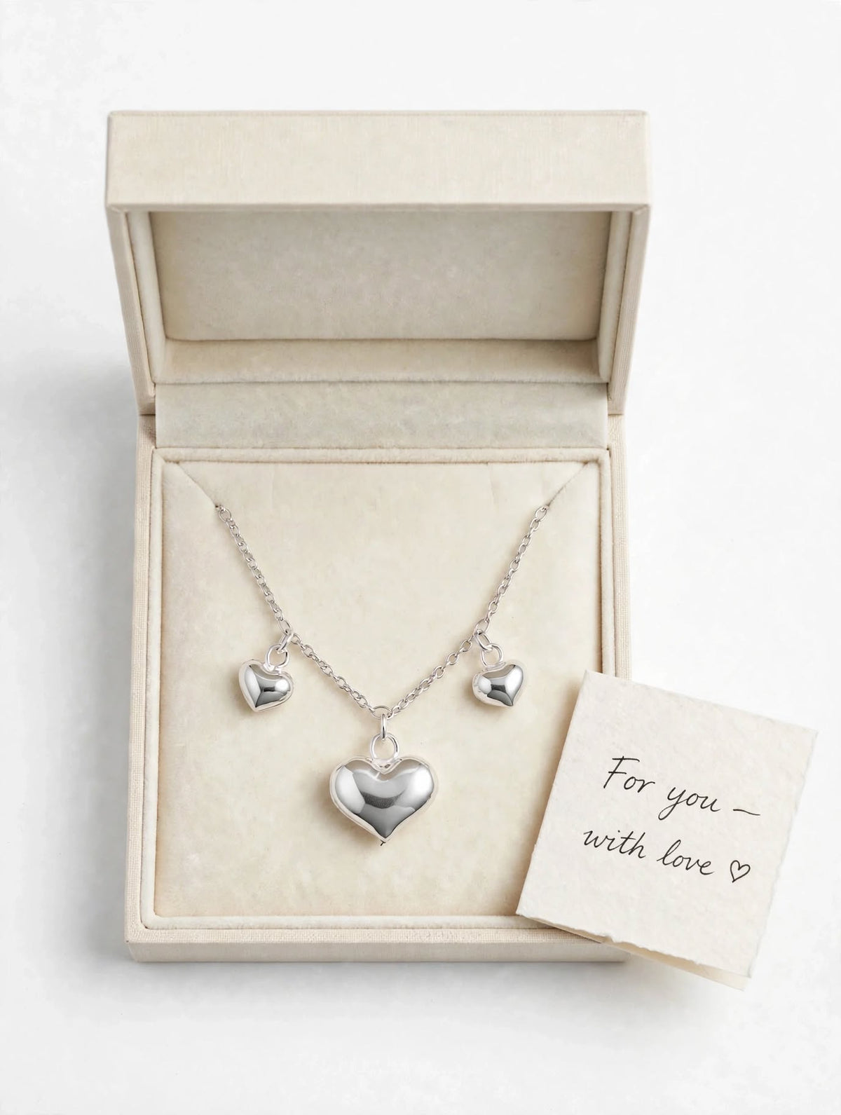 A silver heart pendant necklace with two smaller matching heart charms on a chain, displayed inside an open cream-colored jewelry box. A small handwritten card reading 'For you - with love' rests beside the box.