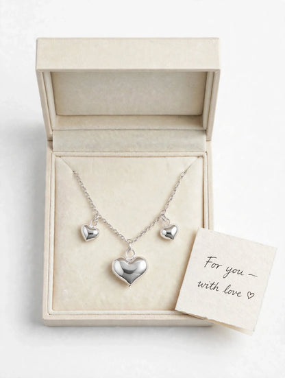 A silver heart pendant necklace with two smaller matching heart charms on a chain, displayed inside an open cream-colored jewelry box. A small handwritten card reading 'For you - with love' rests beside the box.