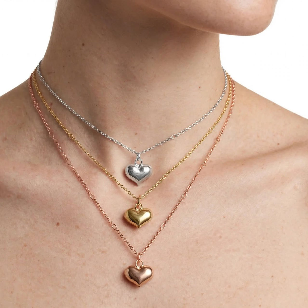 Three delicate heart-shaped pendant necklaces in silver, gold, and rose gold tones layered on a model's neck against a white background.