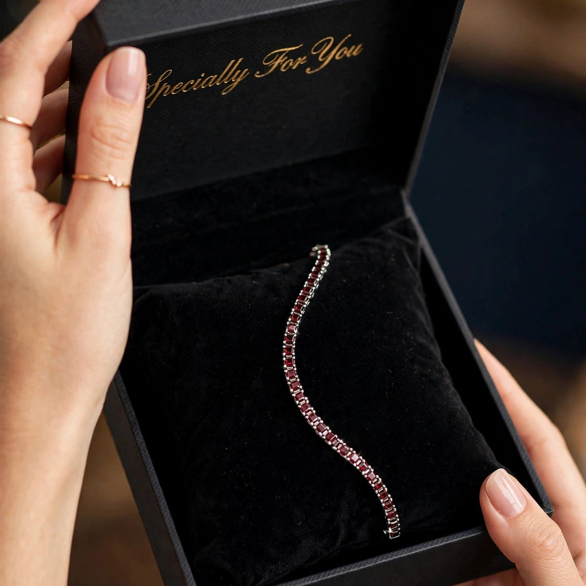 A curved silver tennis bracelet with pink gemstones presented in an open black velvet jewelry box held by two hands.