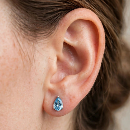 A close-up of a person's ear wearing a small pear-shaped light blue gemstone stud earring in a silver setting.