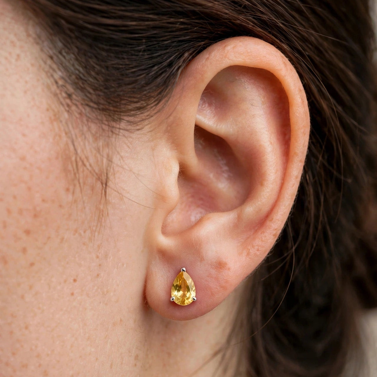 A close-up of a person's ear wearing a small gold teardrop stud earring with a yellow gemstone.