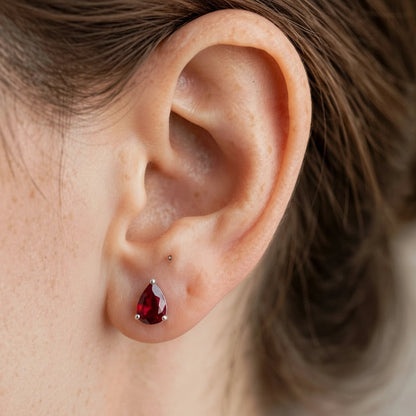 A close-up of a person's ear wearing a small teardrop-shaped dark red gemstone stud earring in a silver setting.