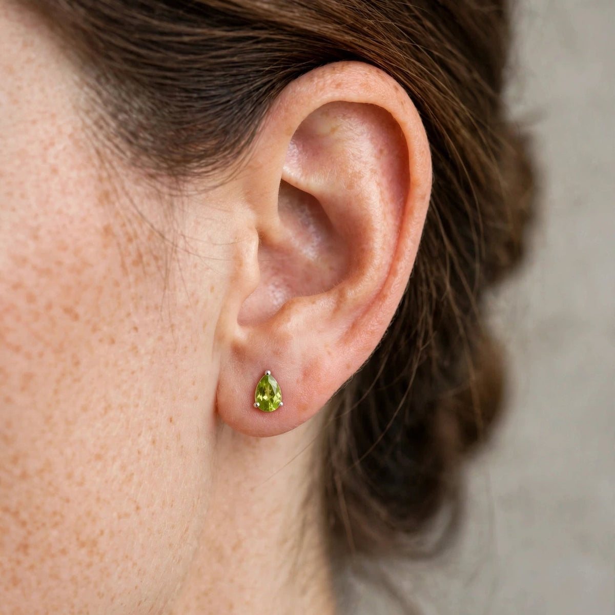 A close-up of a person's ear wearing a small green teardrop-shaped stud earring with a gold setting.