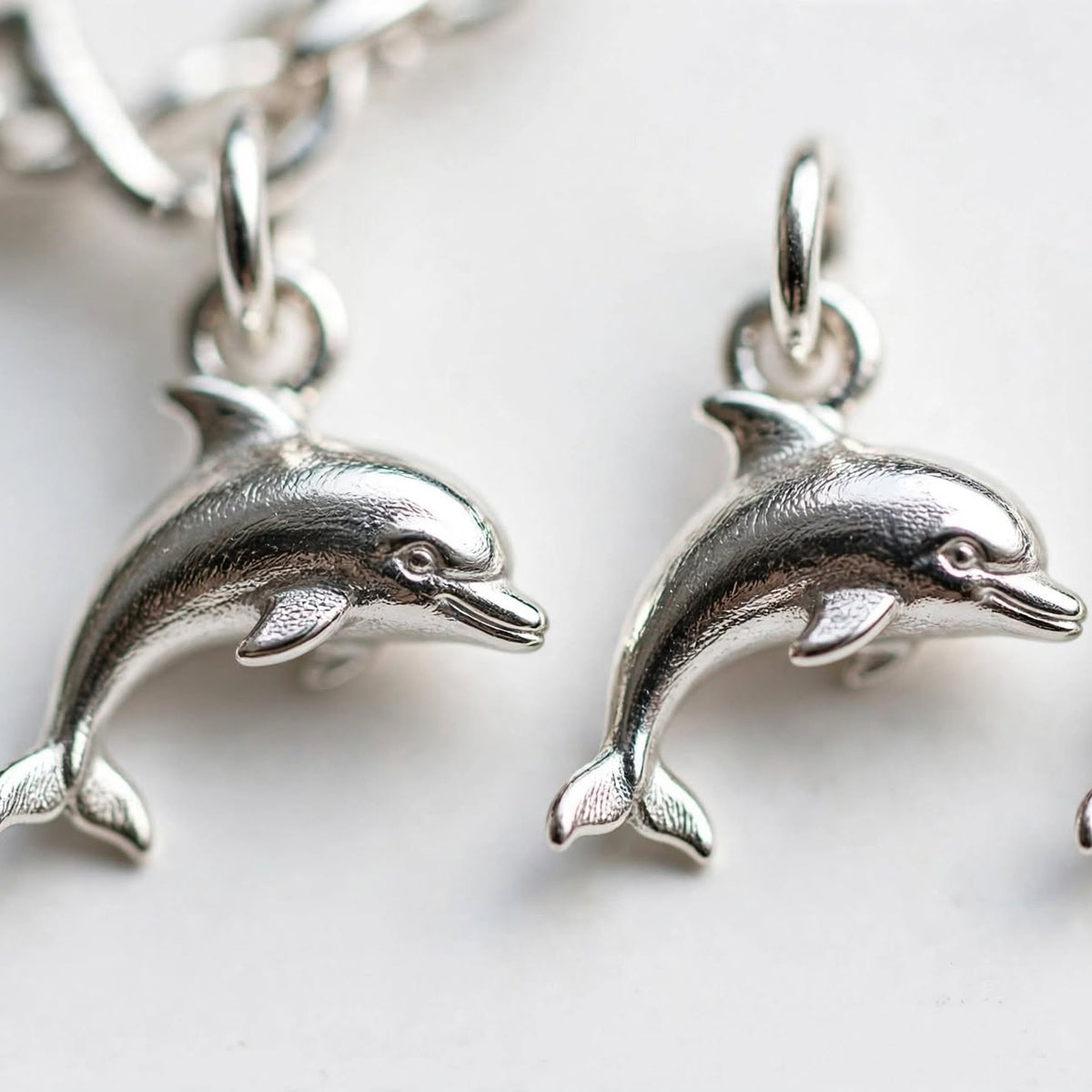 Three silver dolphin charms with attached jump rings, shown on a white background with part of a silver chain visible at the top left.