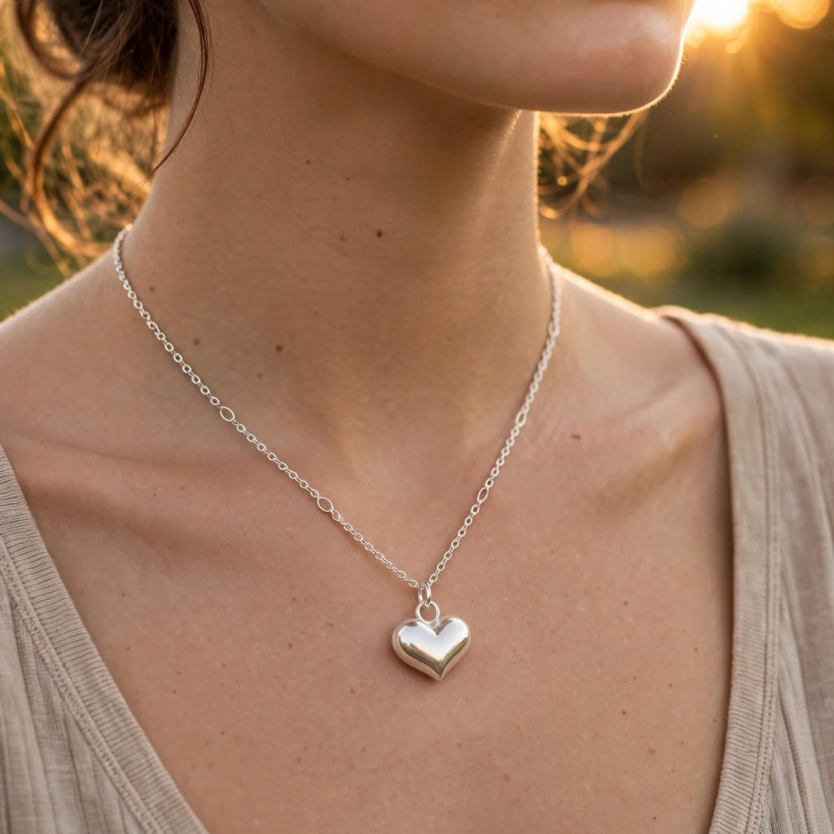 A close-up lifestyle shot of a woman wearing a delicate silver heart pendant necklace on a warm, golden-hour outdoor background.