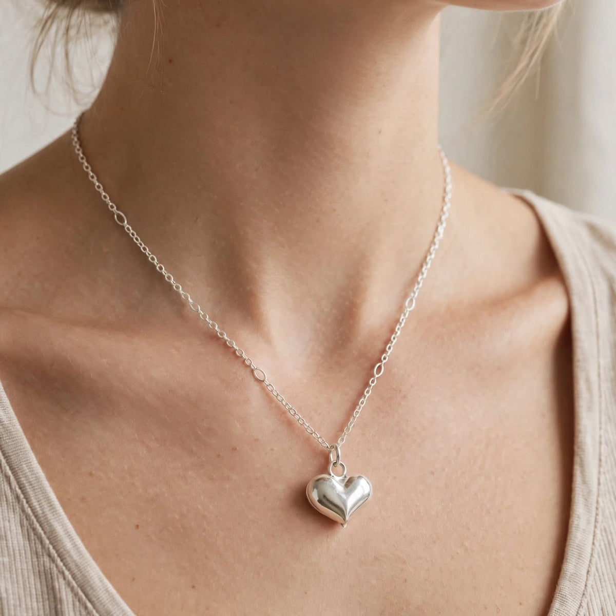 A close-up of a model wearing a delicate silver chain necklace with a polished heart-shaped pendant, paired with a beige ribbed tank top.